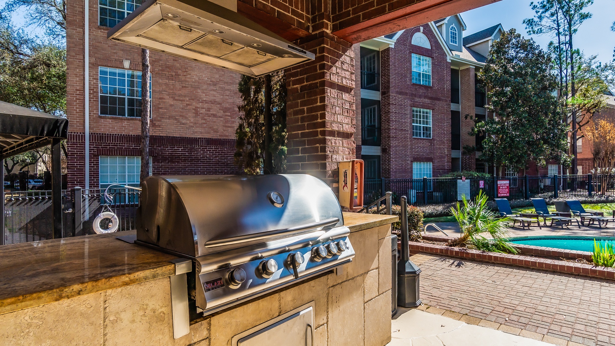 A BBQ grill is on a patio with a pool and trees in the background.